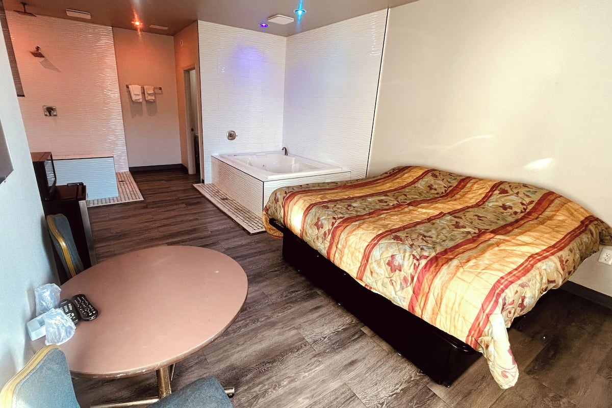 A bedroom with a bed, a small round table, a chair, and a bathroom with a bathtub.