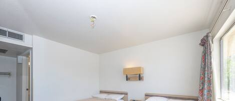 A hotel room with two beds, a bedside table with a lamp, a window with curtains, and a door leading to another room.