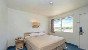 A single bed with a beige bedspread, a bedside table with a phone, a window with curtains, and a door with a lock. - Signature Inn Socorro (Socorro)