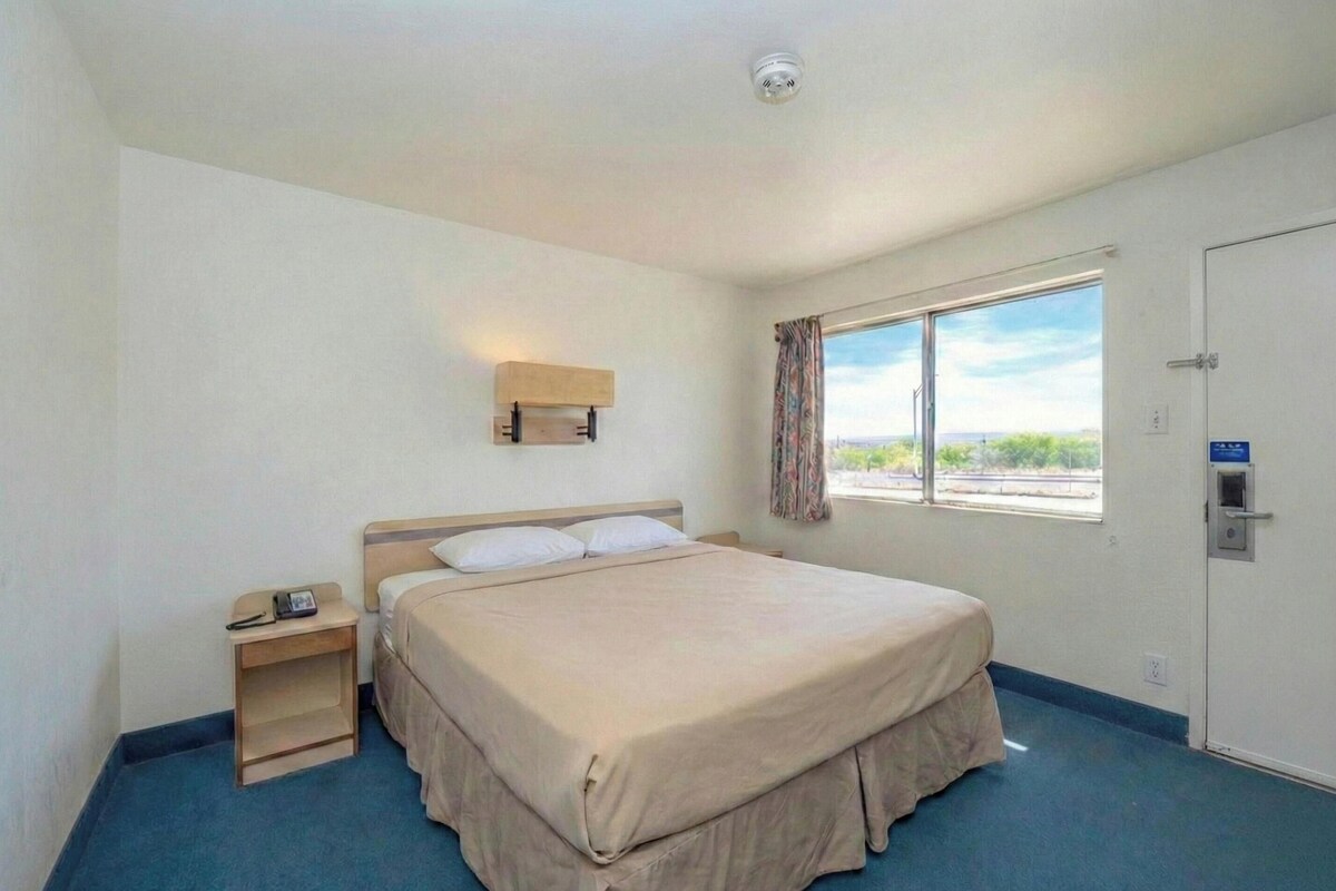 Un lit simple avec une couvre-lit beige, une table de chevet avec un téléphone, une fenêtre avec des rideaux et une porte avec une serrure.