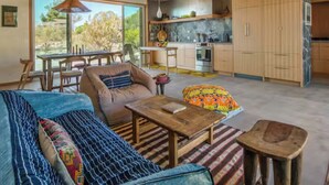 Uma sala de estar ampla com sofá, mesa de centro e mesa de jantar. Há uma cozinha com armários de madeira e uma janela grande que oferece vista para o exterior.