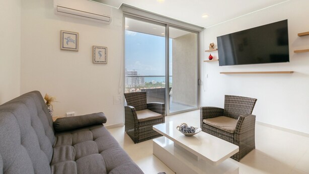 Ein modernes Wohnzimmer mit einer Couch, zwei Korbstühlen, einem Couchtisch, einem Flachbildfernseher und einer Schiebetür zu einem Balkon mit Stadtblick.