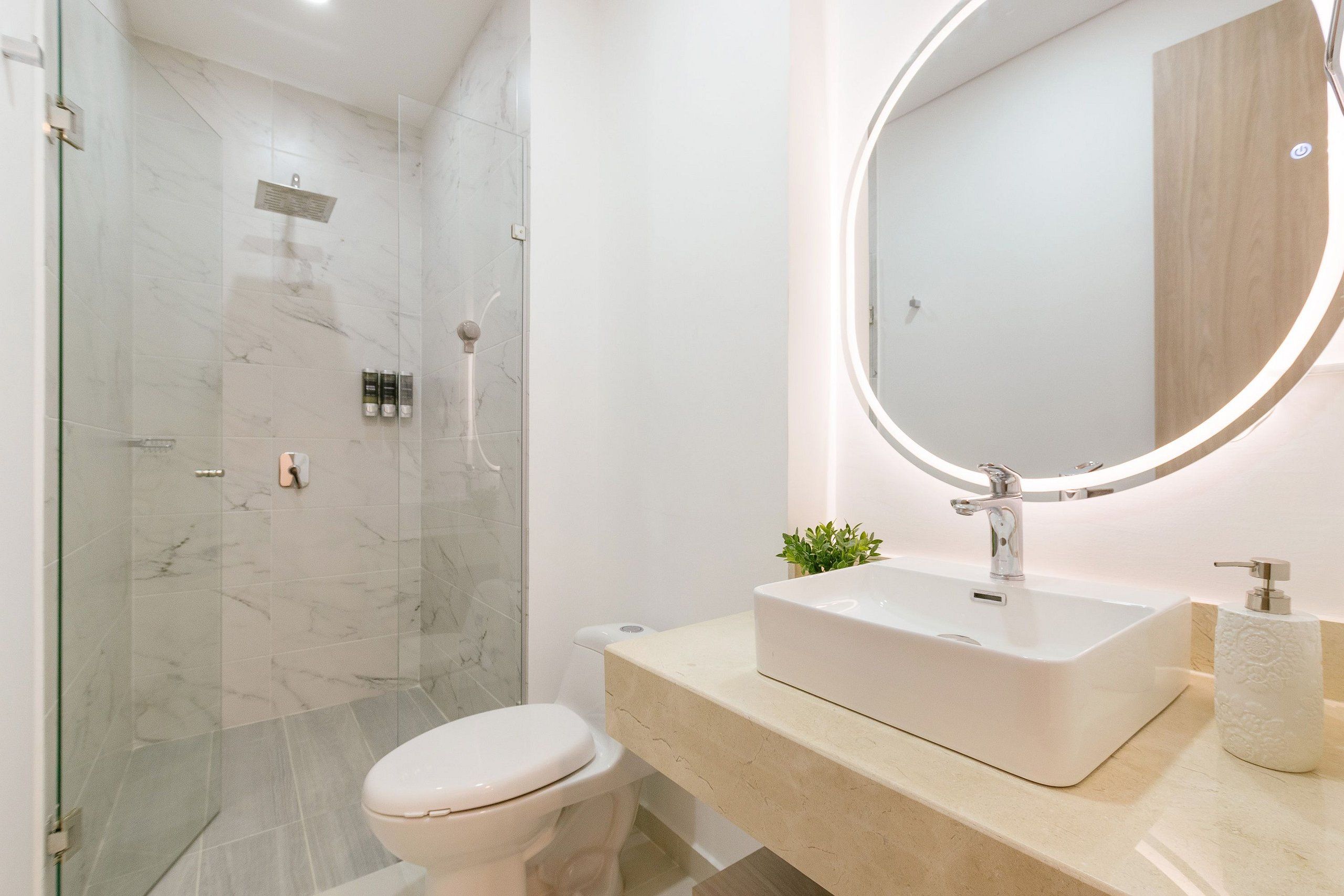 Un baño moderno con cabina de ducha de vidrio, inodoro blanco, tocador color beige con un espejo grande y lavamanos blanco.