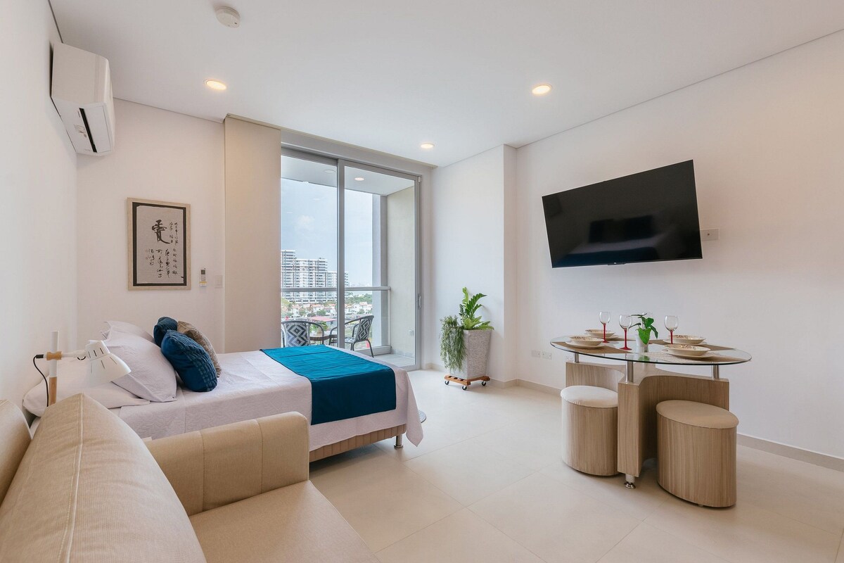 A modern hotel room with a bed, a TV mounted on the wall, a balcony with a view of buildings, and a dining area with a table and chairs.