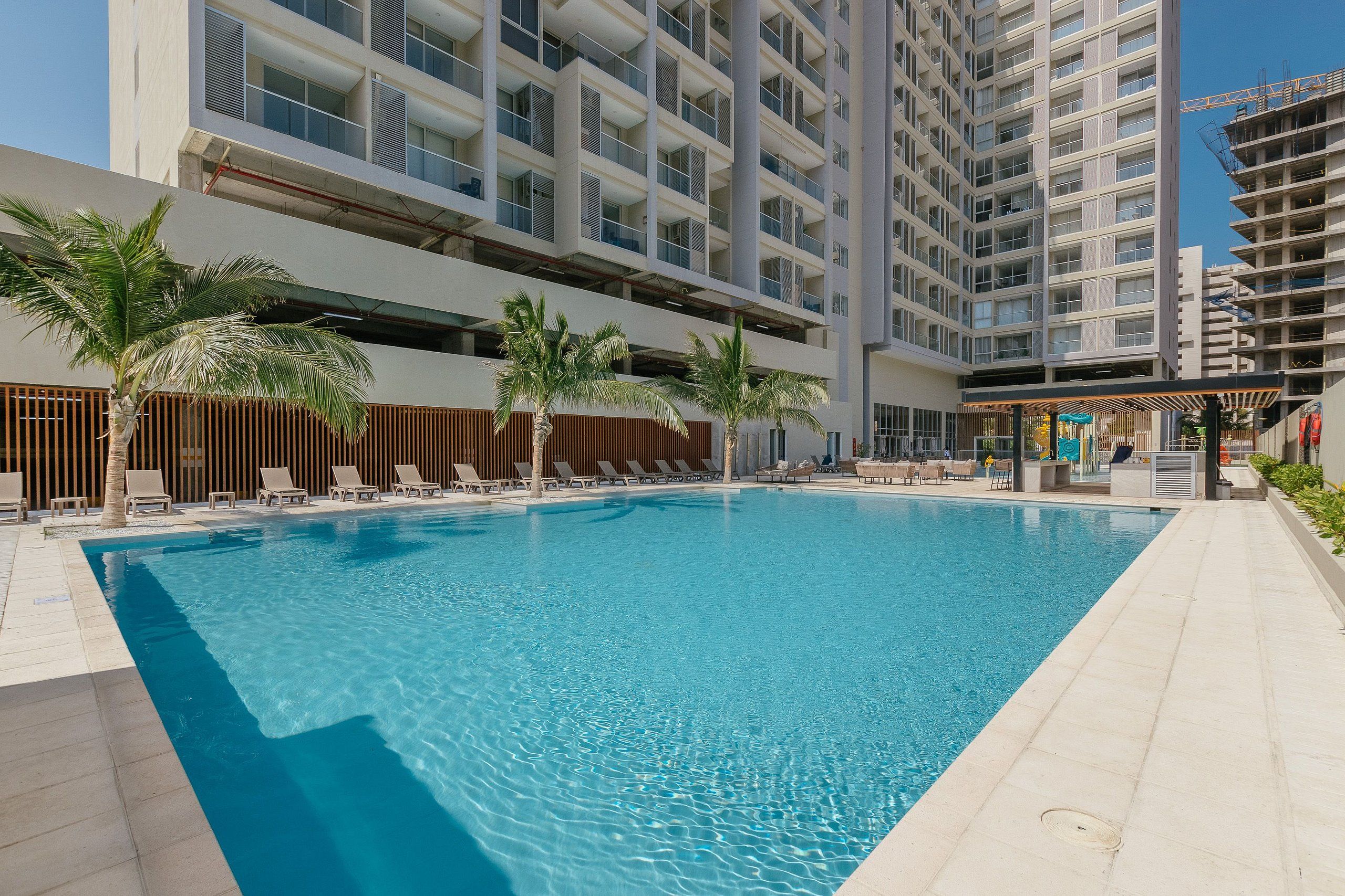 Piscina com espreguiçadeiras e palmeiras, cercada por edifícios modernos.