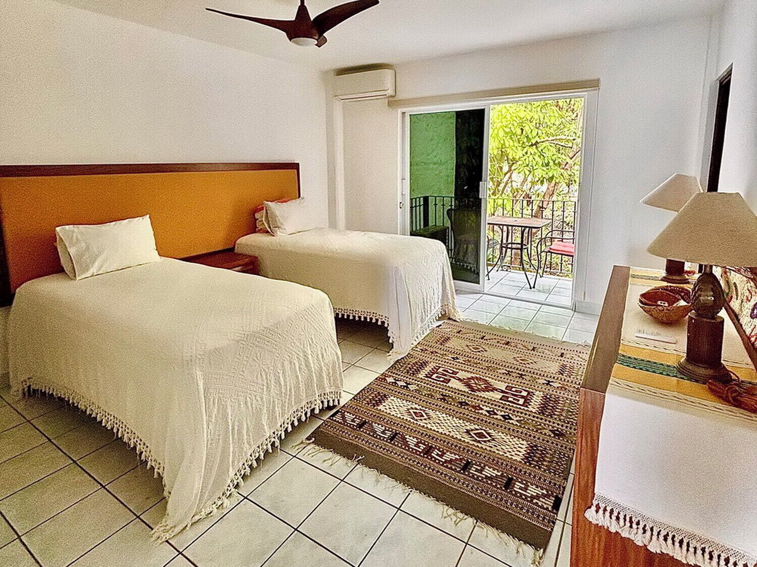Une chambre d’hôtel avec deux lits, un ventilateur de plafond, un balcon avec une table et des chaises, et une lampe sur une table de chevet.