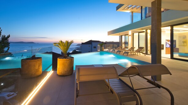 Un espace piscine extérieur moderne, doté de chaises longues, d’une grande plante en pot et offrant une vue sur la mer.