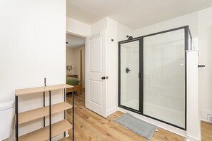 A modern bathroom with a glass shower enclosure, a wooden floor, and a white door. - Queen’s Quarters! 6BR 3B Near Atrium Health & WFU (Winston-Salem)