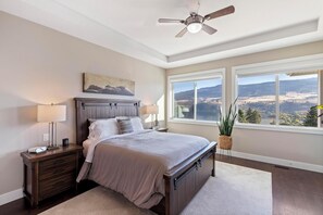 Een slaapkamer met een bed, nachtkastjes, een plafondventilator, een groot raam met uitzicht en een schilderij aan de muur.
