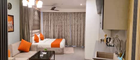 A modern hotel room with a sofa, a bed with orange bedding, a coffee table, a ceiling fan, and a kitchen area with a sink and utensils.