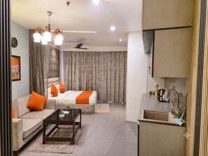 A modern hotel room with a sofa, a bed with orange bedding, a coffee table, a ceiling fan, and a kitchen area with a sink and utensils. - OnlyStay Villa Vista Supernova (Noida)