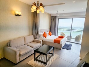 A hotel room with a sofa, a bed with orange pillows, a glass coffee table, a chandelier, and a view of the outdoors. - OnlyStay Villa Vista Supernova (Noida)