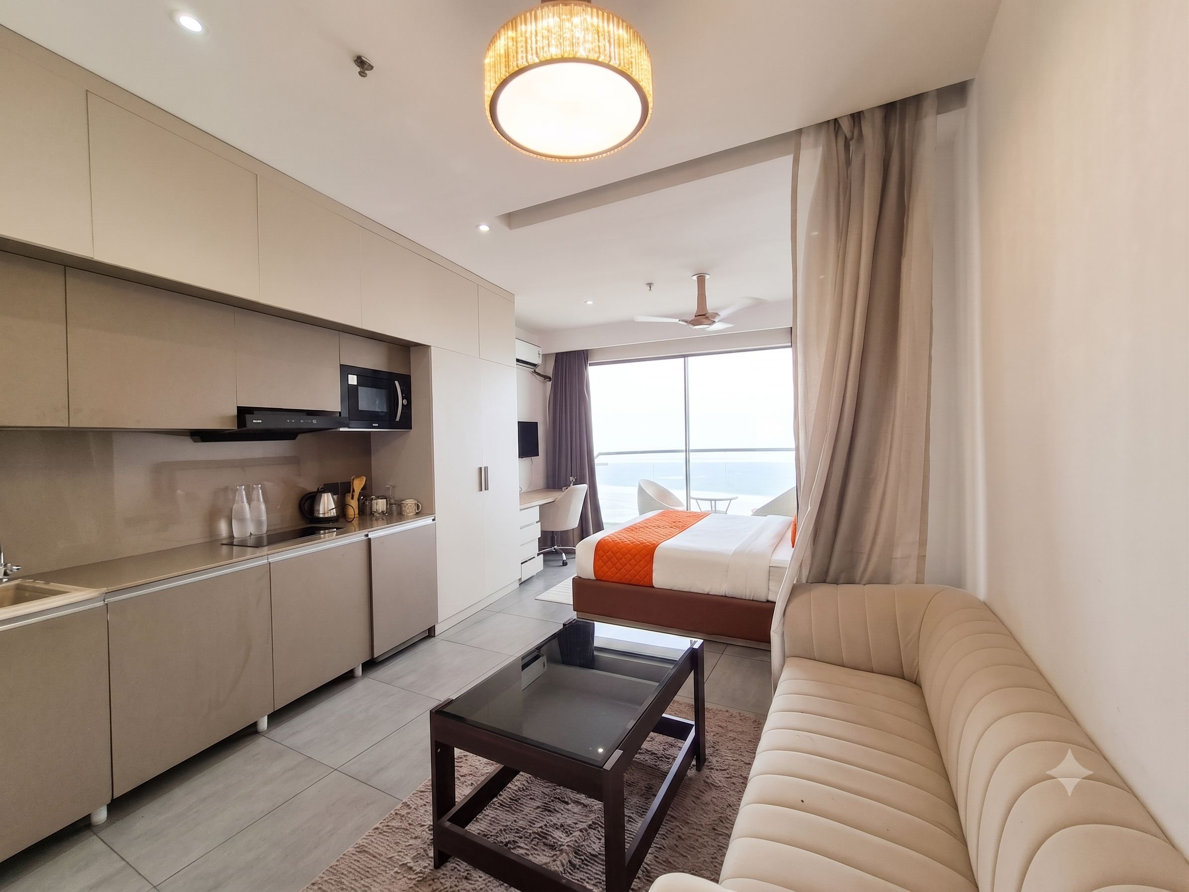 A modern hotel room with a kitchenette, a sofa, a bed, and a large window offering a view.