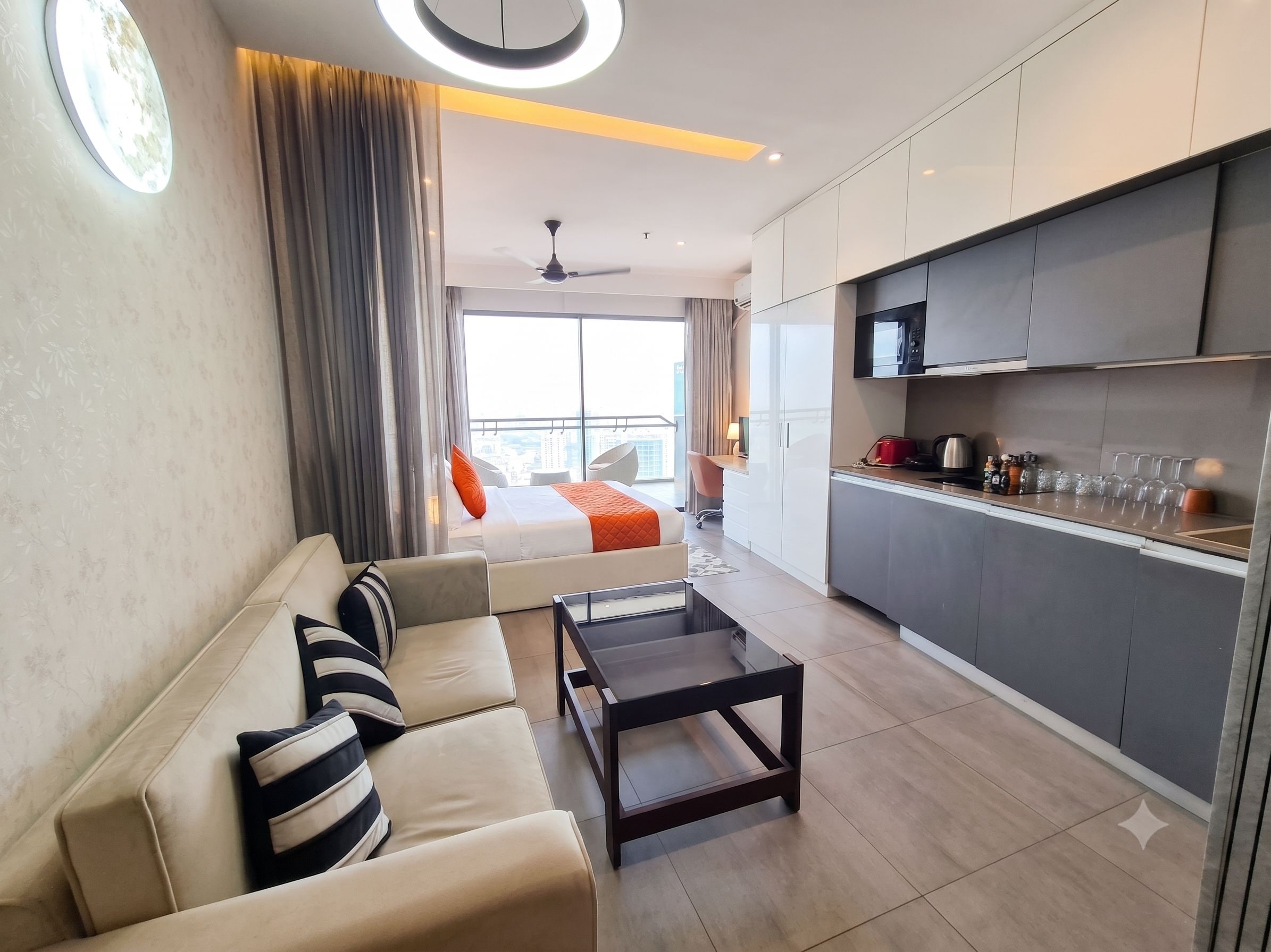 A modern hotel room with a sofa, a coffee table, a bed with an orange blanket, a kitchen area with a microwave, and a view of the city through large windows.