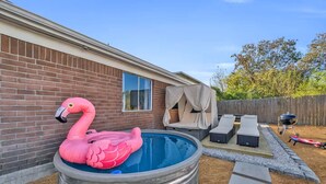 Un giardino sul retro con piscina, un galleggiante a forma di fenicottero, un'area salotto coperta e un barbecue.