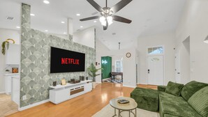 A modern living room with a flat-screen TV, green sofa, wooden flooring, and a ceiling fan. - Poolside Paradise - 3 BR with Game Room & Pool Table! (Ocala)