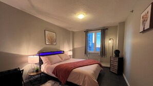 Quarto moderno com cama, abajures, uma pequena mesa, uma cadeira, radiador, janela com vista e luminária no teto.
