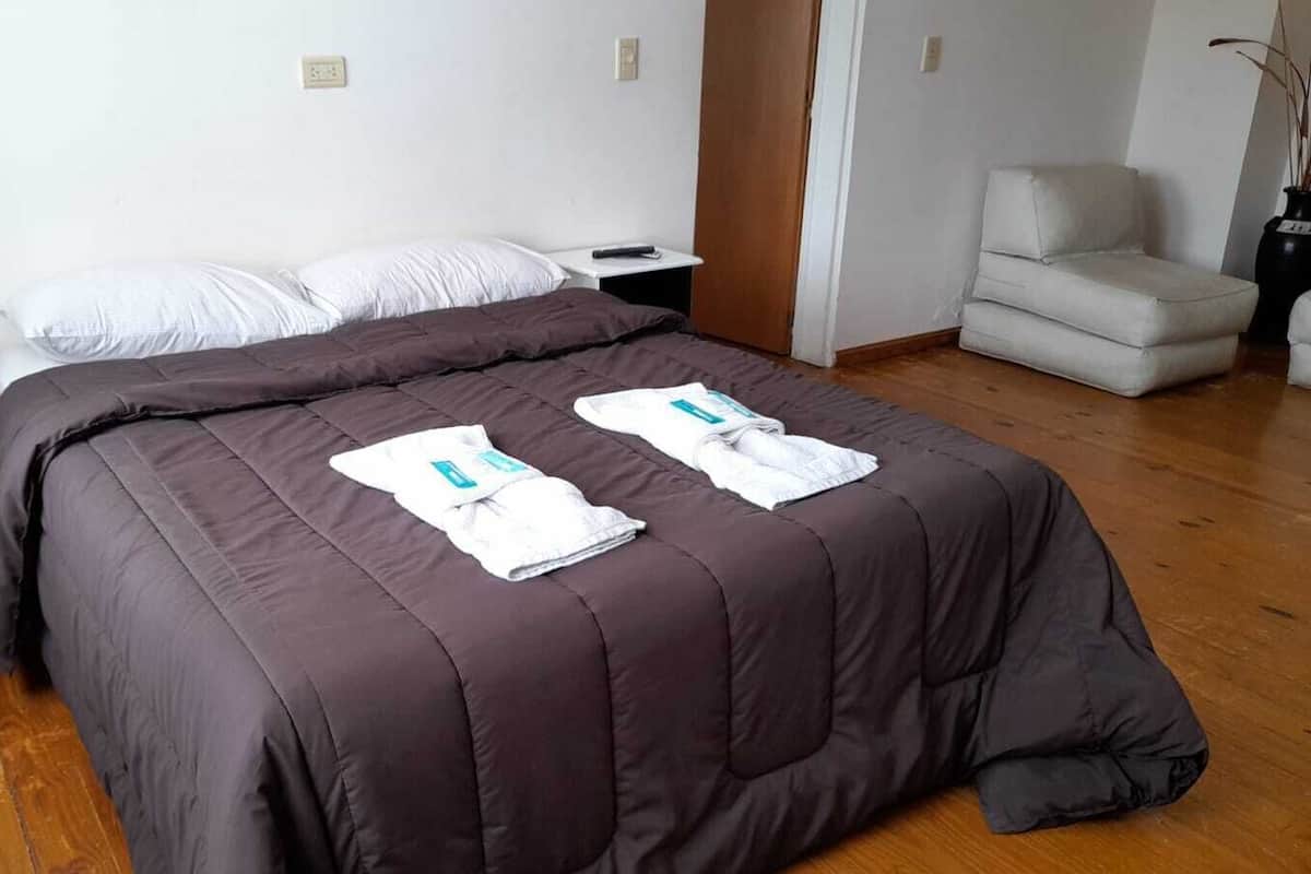 Cama bem arrumada com edredom marrom escuro, duas toalhas brancas e um controle remoto sobre a mesa de cabeceira.