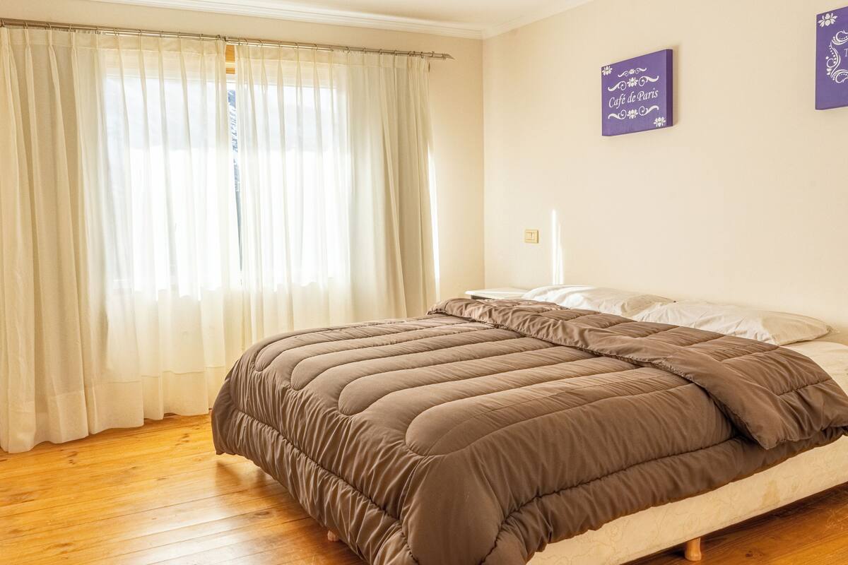 Quarto com cama, piso de madeira, cortinas brancas e parede com um letreiro.