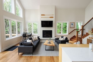 A modern living room with a fireplace, large windows, a staircase, and a dining area.