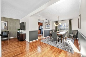 A modern living room with hardwood flooring, a dining area with a table and chairs, a TV mounted on the wall, and a kitchen in the background. - Pool-Fenced Yard-Pool Table-Sleeps 10 (Fairfield)