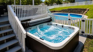 A hot tub on a wooden deck with a white railing, a pool in the background, and a grassy yard.