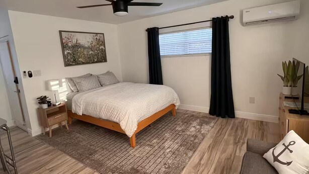 A bedroom with a bed, a ceiling fan, an air conditioner, a window with curtains, a nightstand with a lamp, and a plant. - Cozy Studio - Centrally Located - Free Parking (Orlando)