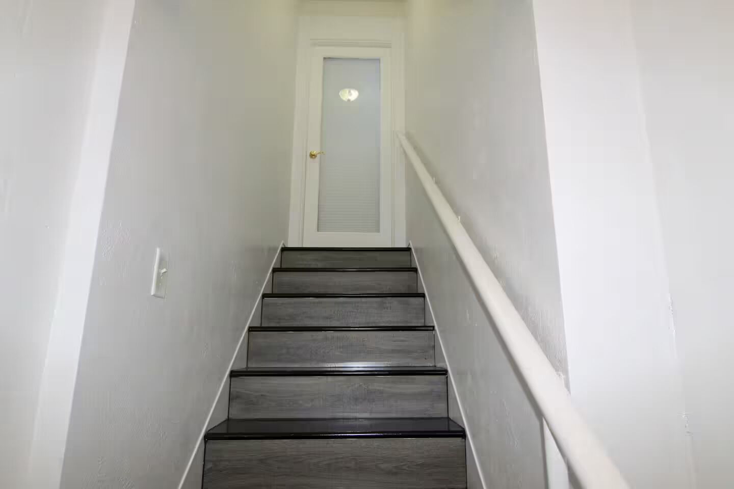 A narrow, white-walled stairway with a wooden handrail and a door at the top.