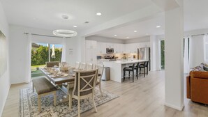 A modern kitchen with white cabinetry, a central island, and a dining area with a table and chairs. - Pluma Verde Vista: 6BR Retreat w/ Pool, Spa, & Games (Coachella)