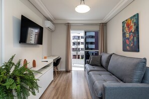 A modern living room with a grey sofa, a white desk, a flat-screen TV, and a large window with curtains.
