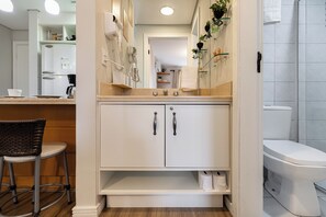 A bathroom with a toilet, sink, and mirror. There is a chair and a shelf with toiletries.