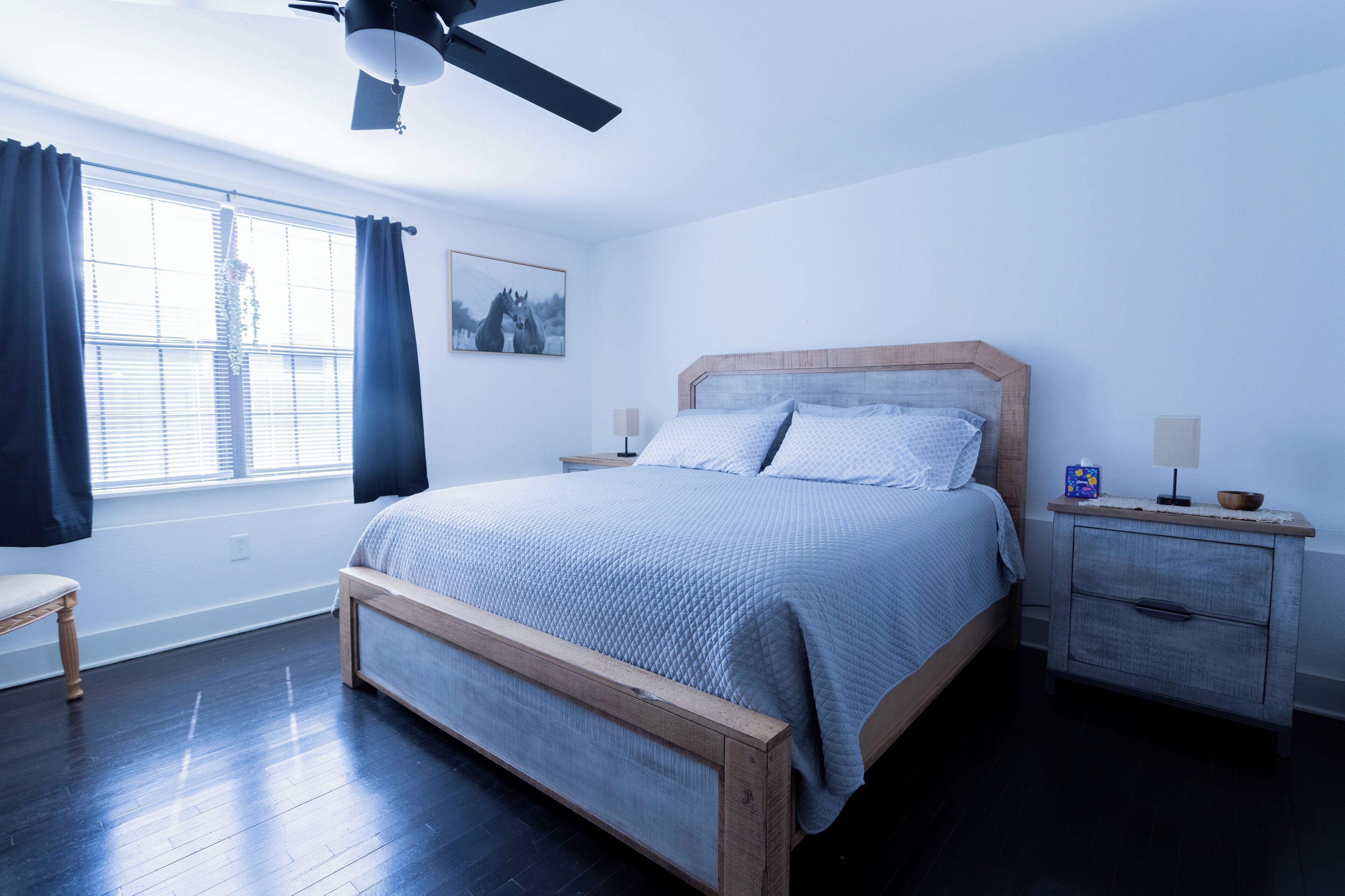 Ein Schlafzimmer mit Bett, Nachttischen, Deckenventilator, Fenster mit Vorhängen und einem Wandbild.