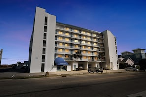 A multi-story hotel with a central entrance, a blue awning, and a parking lot. - Almost Paradise Condo w/ Balcony & Ocean Views (Carolina Beach)