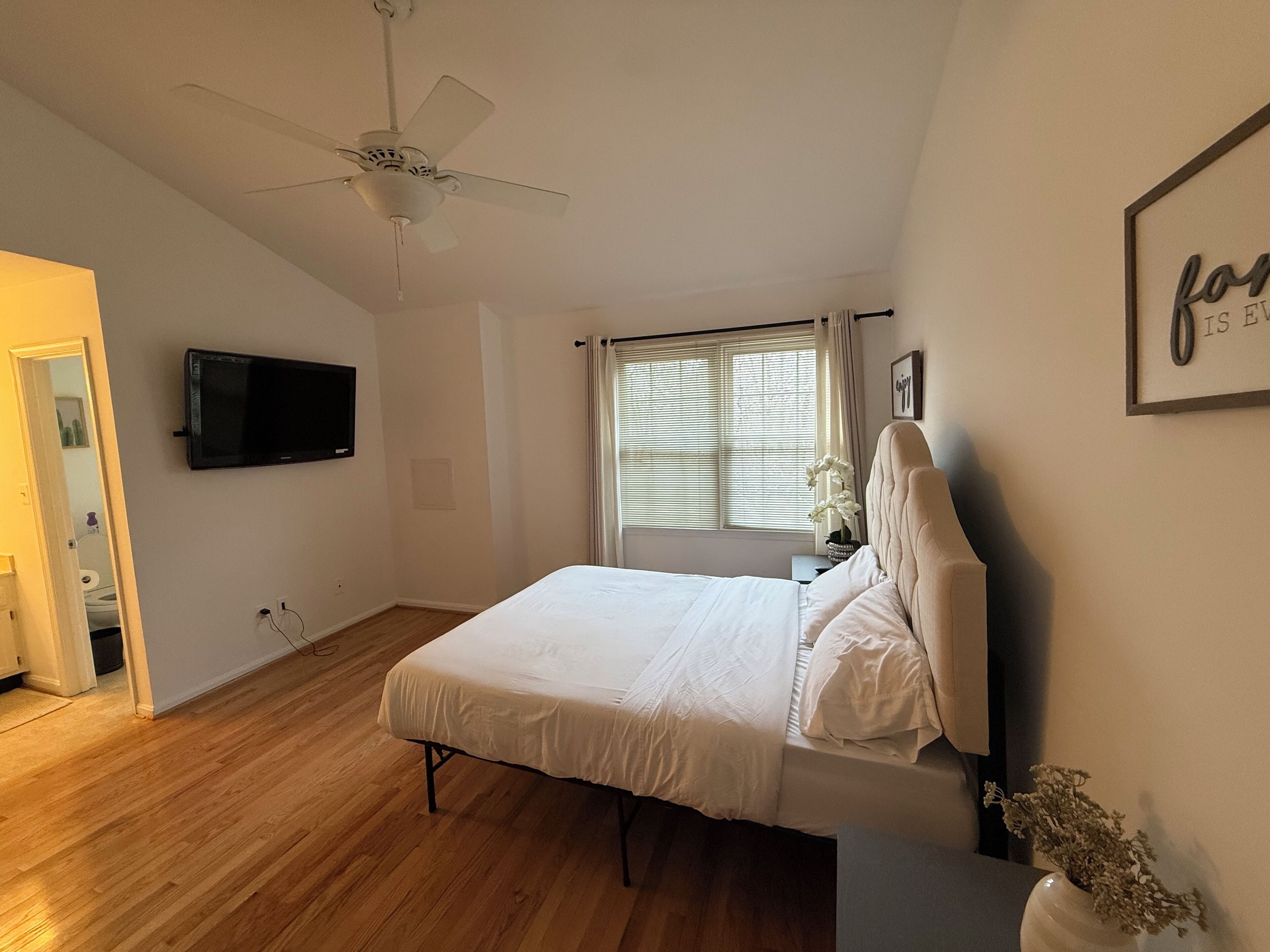 Une chambre à coucher avec un lit, un ventilateur de plafond, une télévision et une fenêtre avec des rideaux.