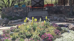 Ein überdachter Pavillon mit einer Steinsäule und Stufen, umgeben von bunten Blumen, mit einer Berglandschaft im Hintergrund.