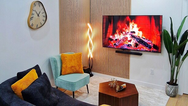 Ein modernes Wohnzimmer mit einer gebogenen schwarzen Couch, einem türkisfarbenen Sessel, einer Wand aus Holzlamellen, einem Flachbildfernseher, einer Wanduhr und einer Topfpflanze.