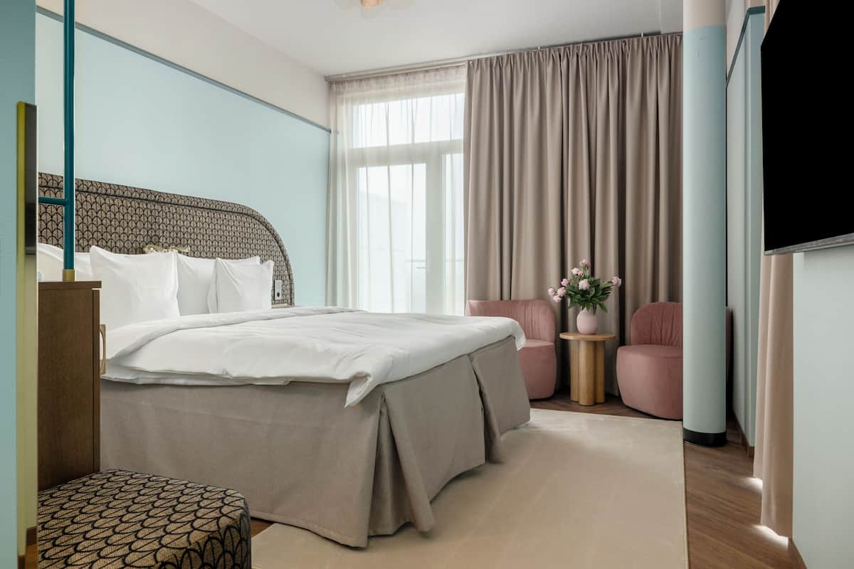 A hotel room with a bed, a small seating area, a TV, and a window with curtains.