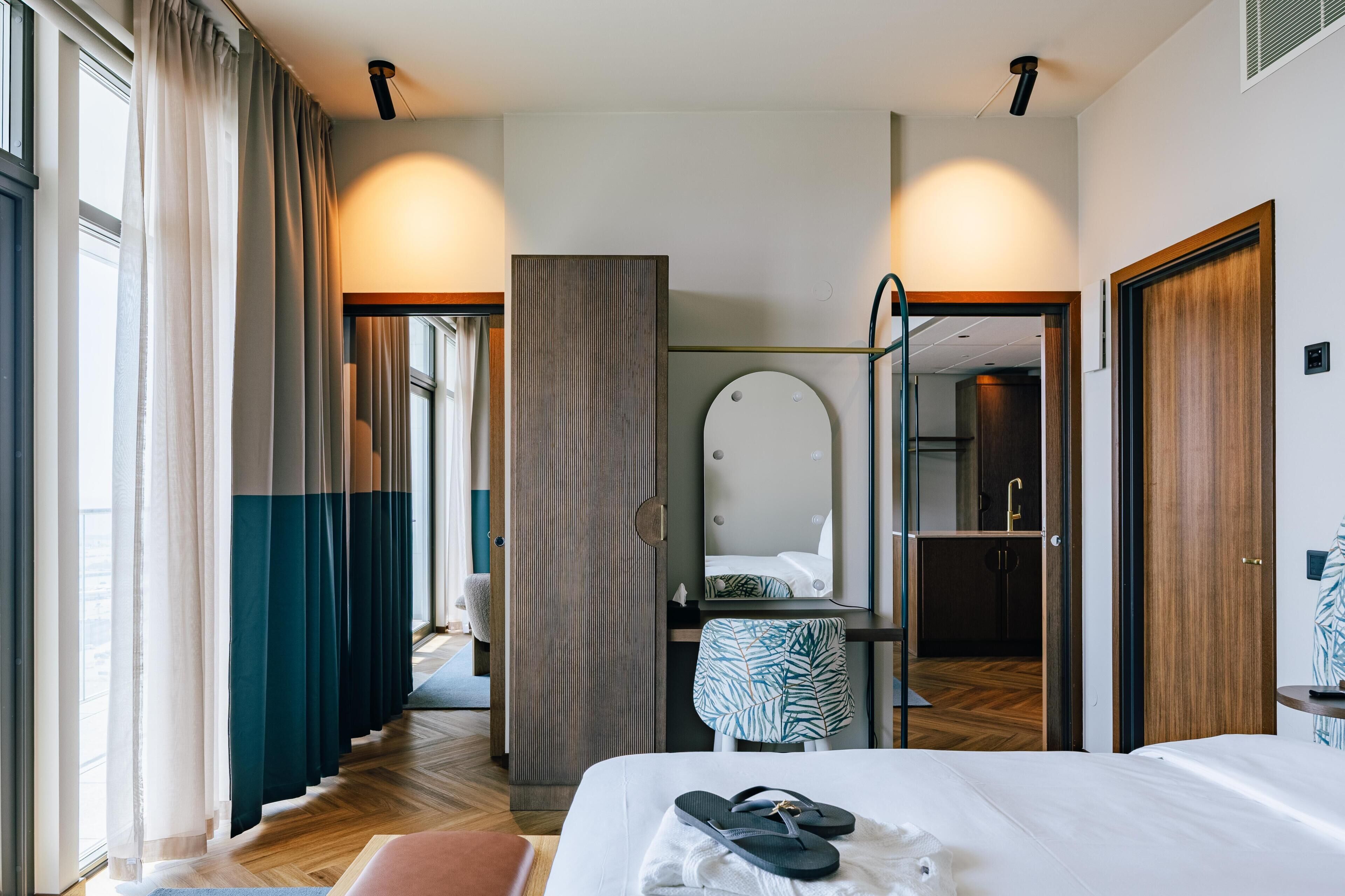 Ett hotellrum med en säng, en spegel, ett badrum med handfat och utsikt över korridoren.