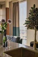 Una habitación con vistas a la ciudad, una ventana con cortinas, una planta, una botella de vino y dos copas sobre una mesa.