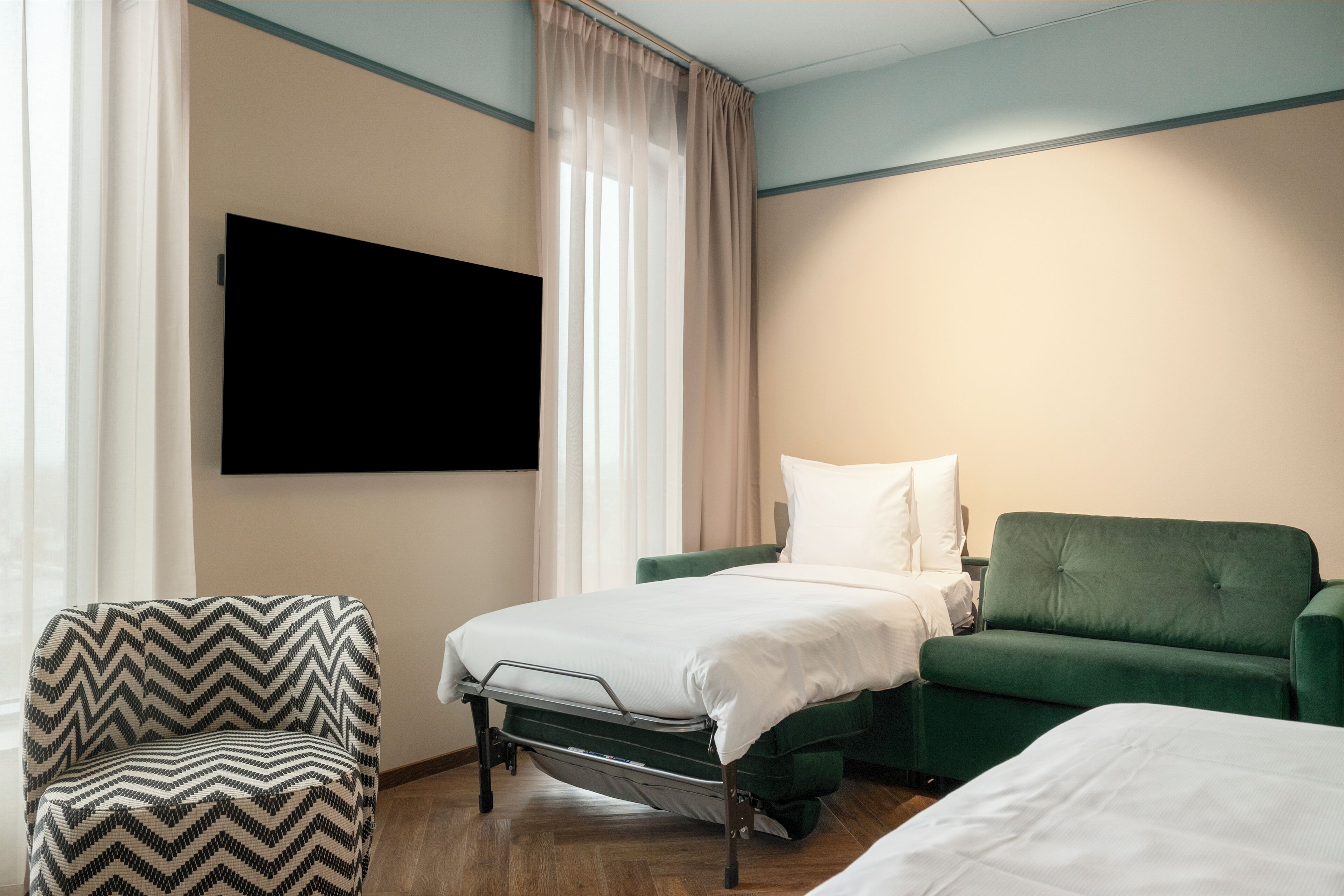 A hotel room with a bed, a green sofa, a chair, and a TV mounted on the wall.