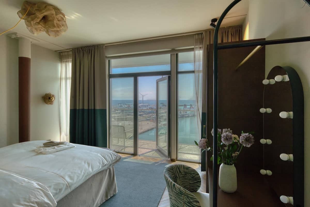 A hotel room with a bed, a chair, a small table, a vase with flowers, and a large window with a view of the city.