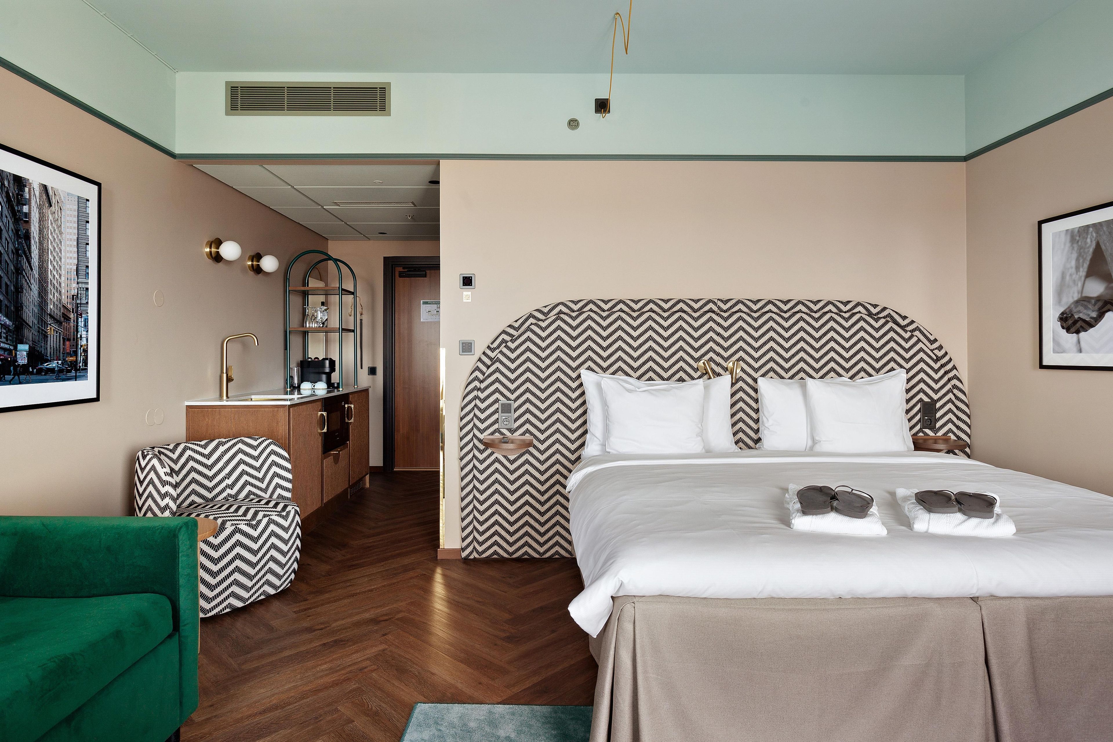 Una habitación de hotel con una cama grande, un sillón verde, un escritorio pequeño con un espejo y un cuadro en la pared.
