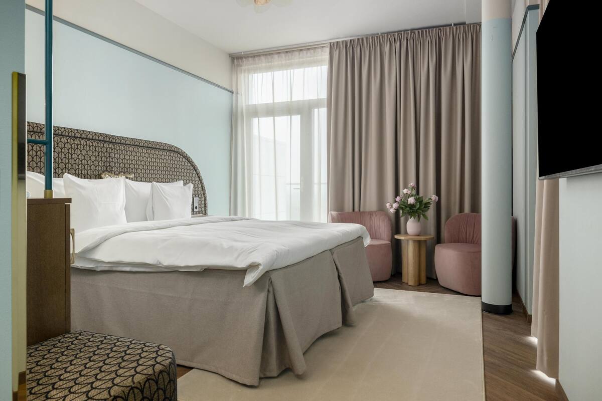 Une chambre d’hôtel avec un lit, un petit coin salon, une télévision et une fenêtre avec des rideaux.