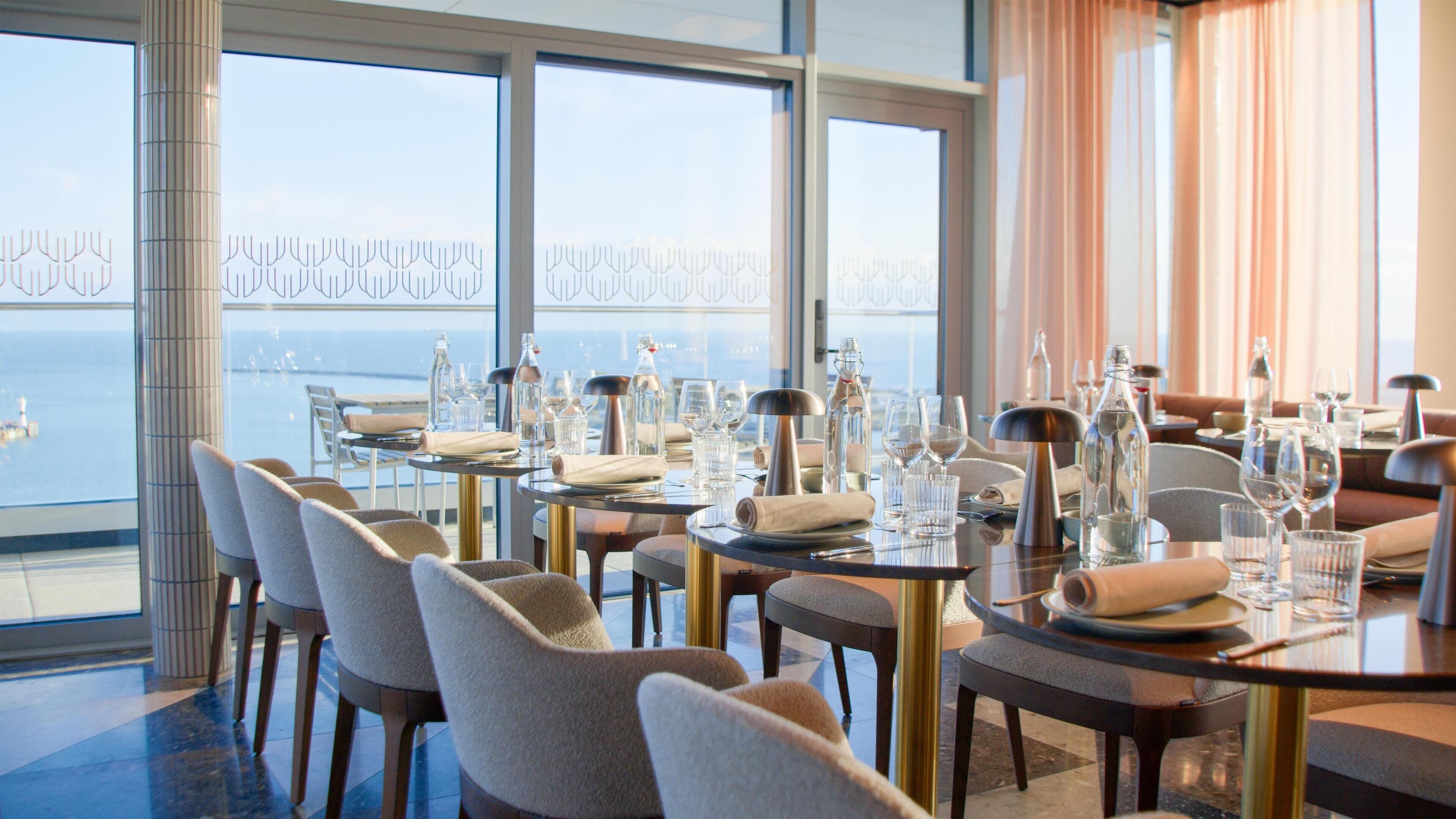 A dining area with tables set for service, large windows with a view of the sea, and a high ceiling.