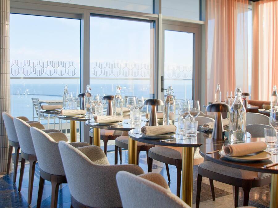 A dining area with tables set for service, large windows with a view of the sea, and a high ceiling.