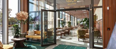 Un espace d’accueil d’hôtel au design moderne, comprenant un comptoir d’accueil, un coin salon et de grandes fenêtres laissant entrer la lumière naturelle.