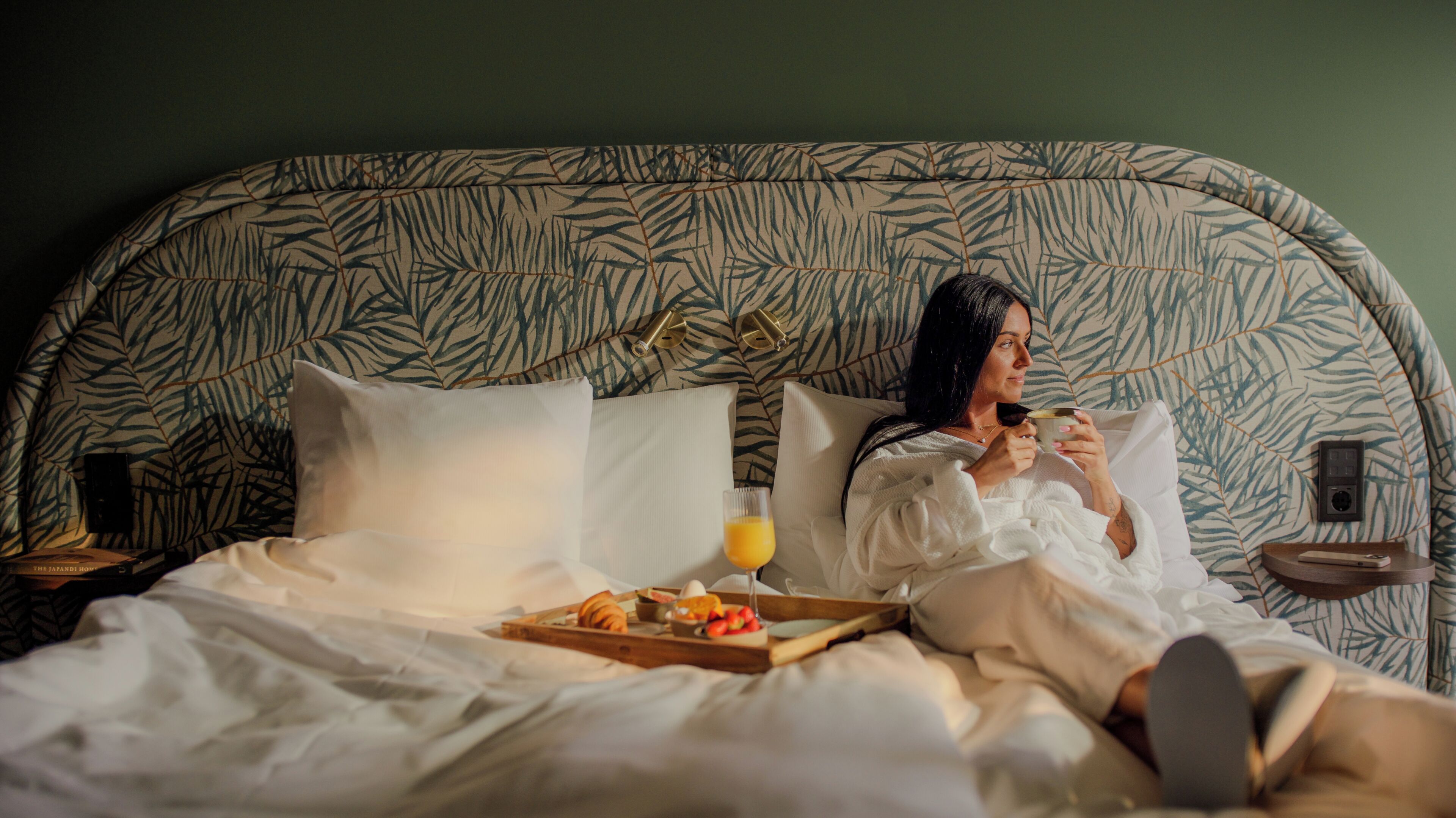 Een persoon in bed met een dienblad ontbijt, een nachtkastje met een lamp en een hoofdeinde met een patroon.
