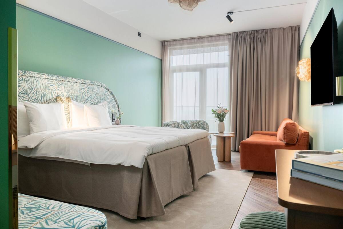 A hotel room with a bed, a desk, a chair, a window with curtains, and a TV mounted on the wall.