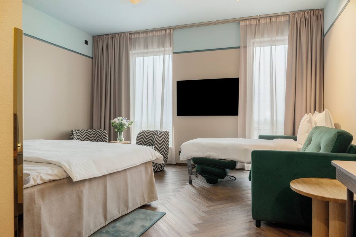 A hotel room with two beds, a green armchair, a small table, and a flat-screen TV.