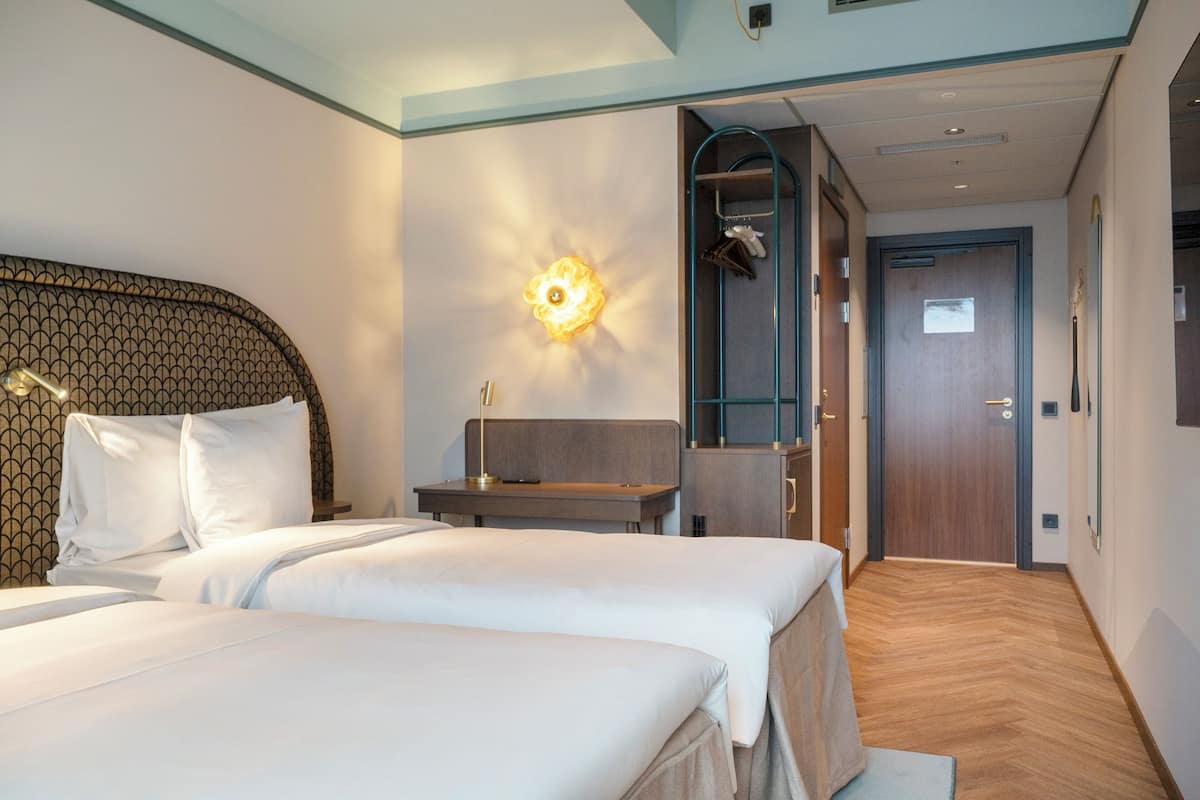 A hotel room with two beds, a nightstand, a wall-mounted lamp, a wooden door, and a mirror.